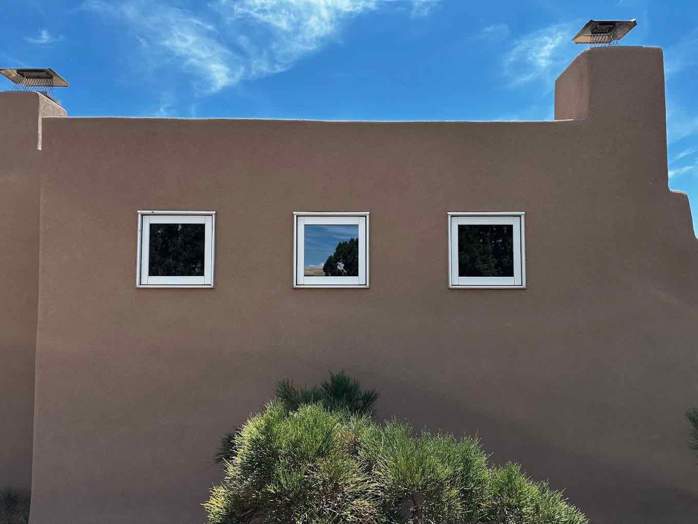 Three square windows set into a tall adobe wall under a deep blue Santa Fe sky, the glass clean enough to mirror it.